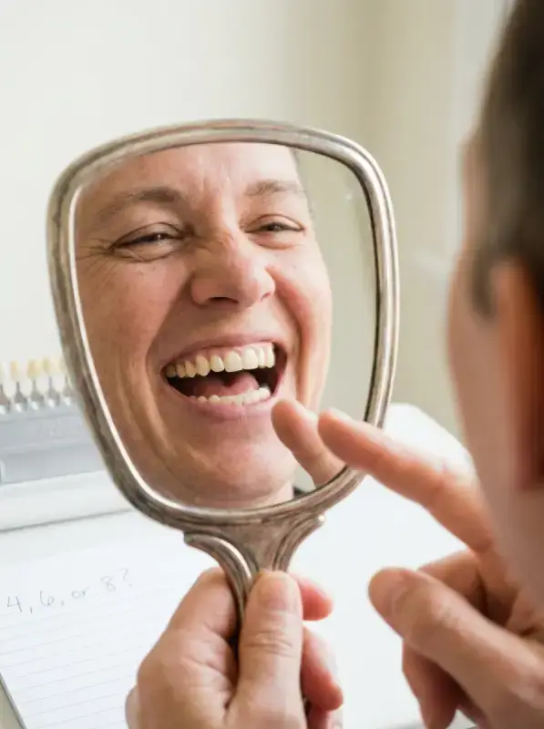 Close-up of a hand mirror held at eye level showing a full laughing smile with the canines and first premolars visible, used to count teeth before choosing four, six, or eight veneers
